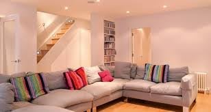 Modern living room with beige L-shaped sectional sofa, colorful throw pillows, wooden staircase, and built-in bookshelves