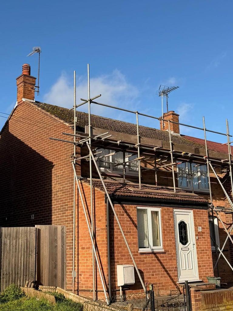 Red brick house under renovation with metal scaffolding, two chimneys, and TV antenna visible against blue sky