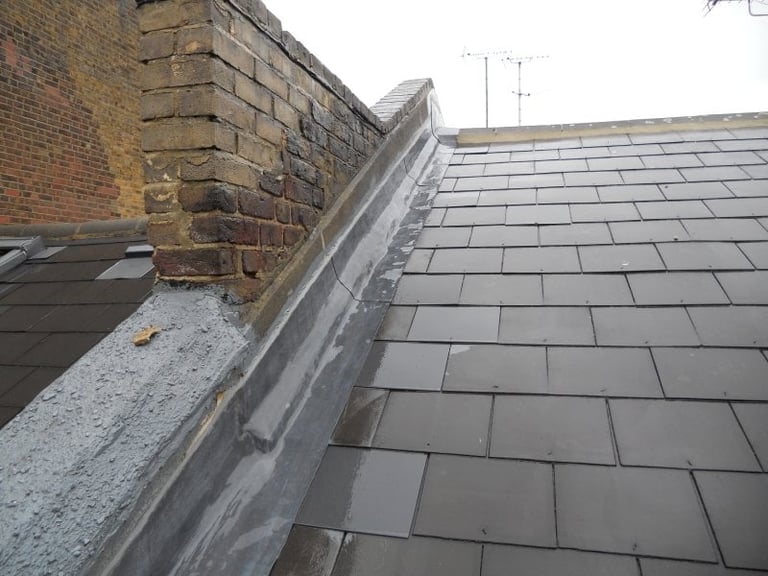 Close-up of roof edge showing slate tiles, metal gutter, brick chimney, and TV antenna against sky