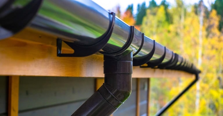 Black metal gutter and downspout on yellow house with autumn forest trees in background