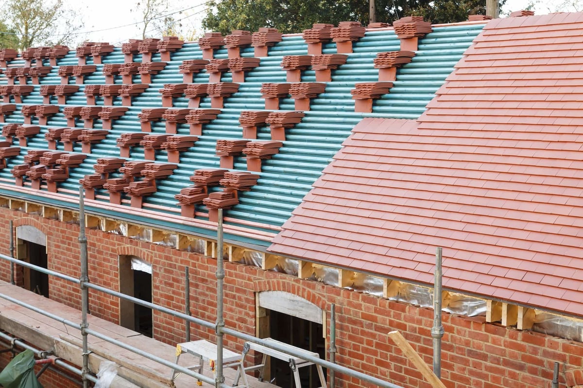 Roof under construction with turquoise and red metal sheets, brick building walls, and yellow safety railings visible below