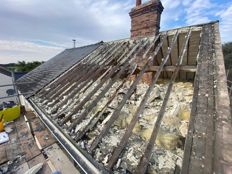 Exposed wooden roof structure with missing tiles and deteriorated underlayment, brick chimney visible against cloudy sky