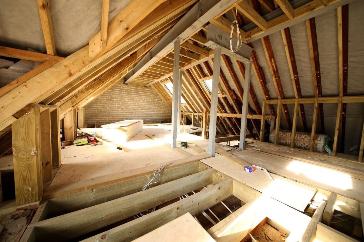 Unfinished attic interior with exposed wooden beams, rafters, and joists during construction, showing insulation and flooring work in progress