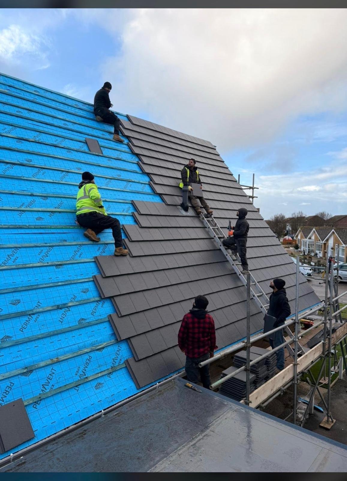 Construction workers installing dark roof tiles on a residential building, with blue underlayment visible and scaffolding surrounding the structure