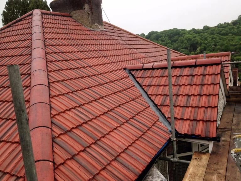 Angled view of red clay tile roof with metal framework and gutters against forested hillside background