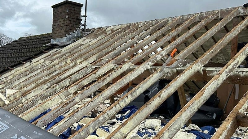 Damaged roof with exposed wooden beams and debris scattered across it, showing deterioration with visible blue tarp and stone chimney in background